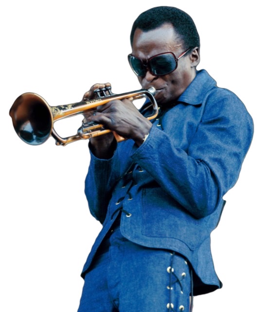 Miles Davis in a studded blue jean jacket and pants blowing on the trumpet.