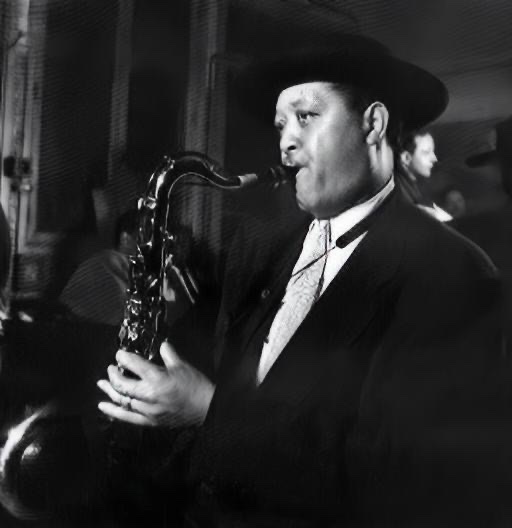 A black and white photograph of Lester Young playing his saxophone while facing in left profile.