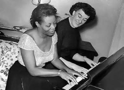 A black and white photograph of Mary Lou Williams and Marian McPartland sitting on the same piano bench during the first episode of NPR's program "Piano Jazz" in 1978.