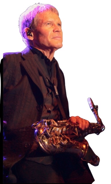 An enhanced photographic cutout of David Sanborn at age seventy-one sitting down wearing a brown striped jacket holding his alto saxophone in his lap while sitting in a chair looking up to his left.