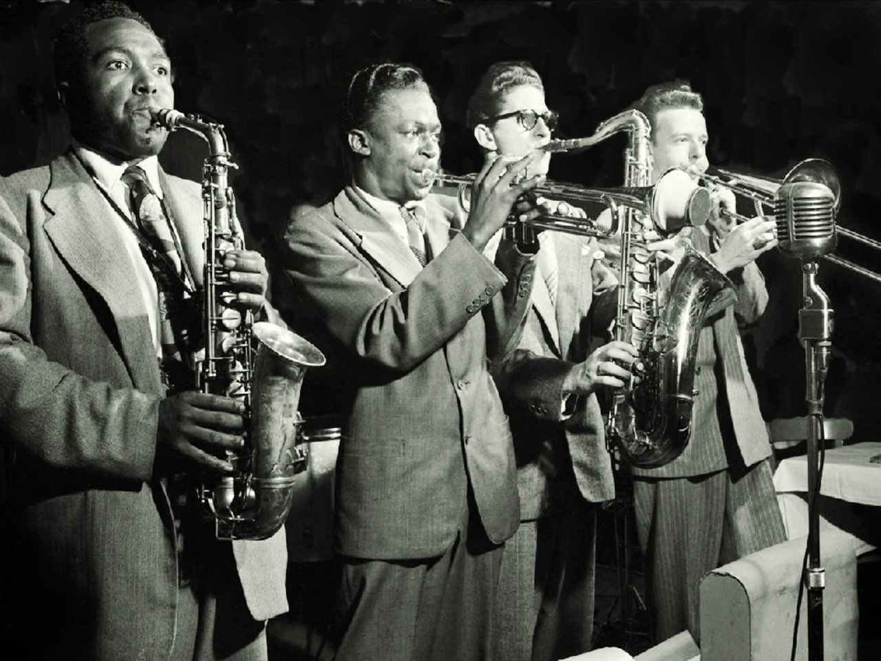 A black and white photograph by Herman Leonard of from left to right Charlie Parker, Miles Davis, Allen Eager, and Kai Winding respectively playing the alto saxophone, trumpet with mute, tenor saxophone, and trombone.