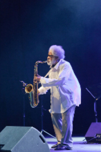 A color photograph of Sonny Rollins with white bushy hair and beard wearing a free flowing white shirt and blue pants👖while playing his saxophone on stage at a microphone.