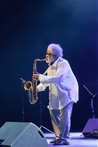 An enhanced color photograph of Sonny Rollins with white bushy hair and beard wearing a free flowing white shirt and blue pants👖while playing his saxophone on stage at a microphone.