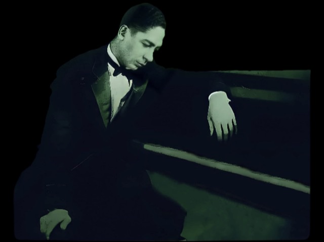 Jelly Roll Morton in his twenties wearing a tuxedo at piano turned sideways with his head bowed.