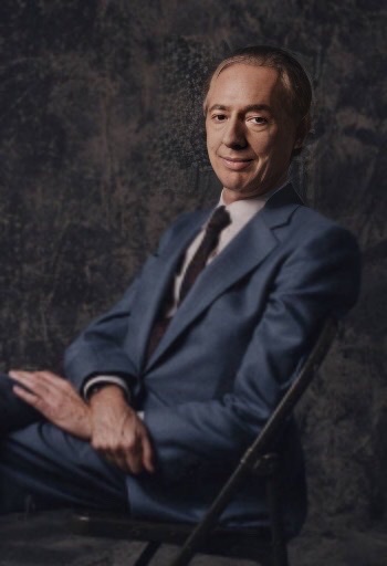 An enhanced color photograph of Leonard Feather with a wry half-smile sitting in a chair 🪑 with hands crossed on his lap facing left wearing a blue suit.