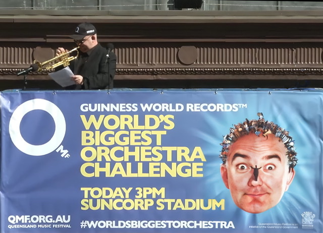 Conductor James Morrison playing his trumpet while conducting the World's Largest Orchestra with its banner displayed underneath him.