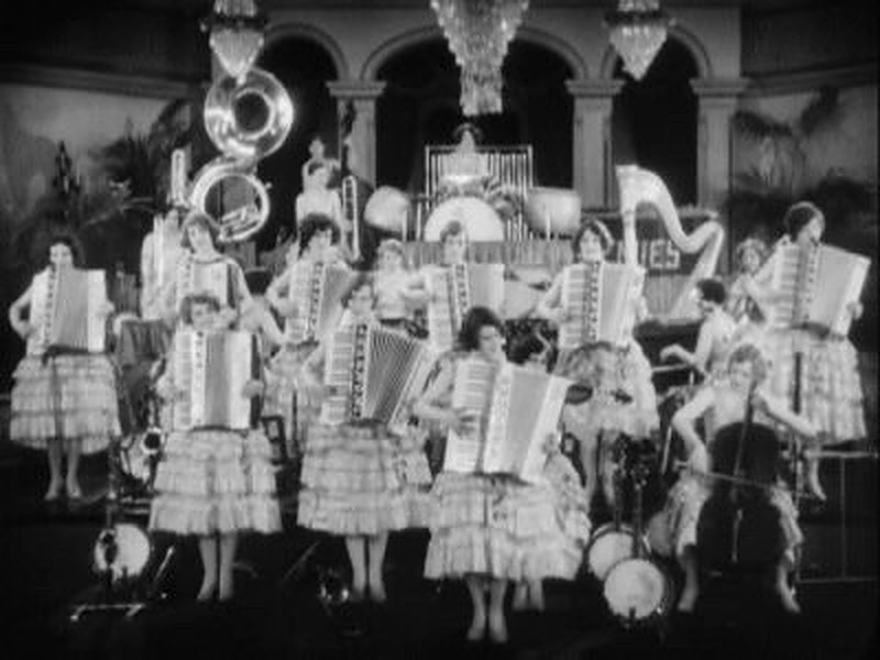 A blurry black and white photograph of The Ingenues with nine accordions and a cello on far right.