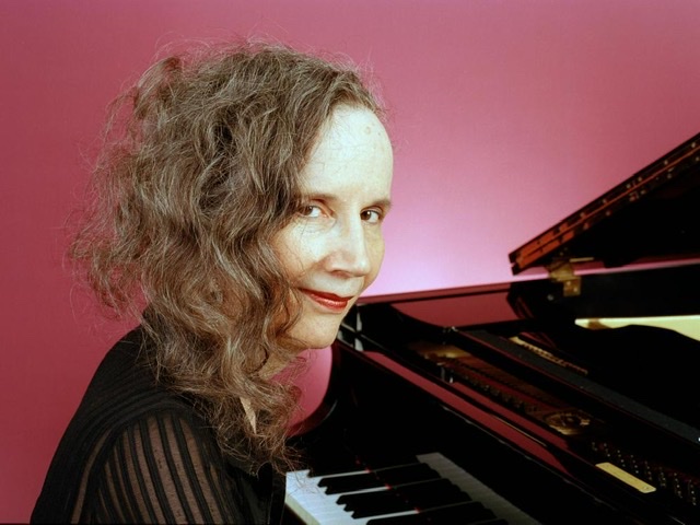 A color photograph of an older Joanne Brackeen sitting at piano with head turned towards camera revealing a high forehead