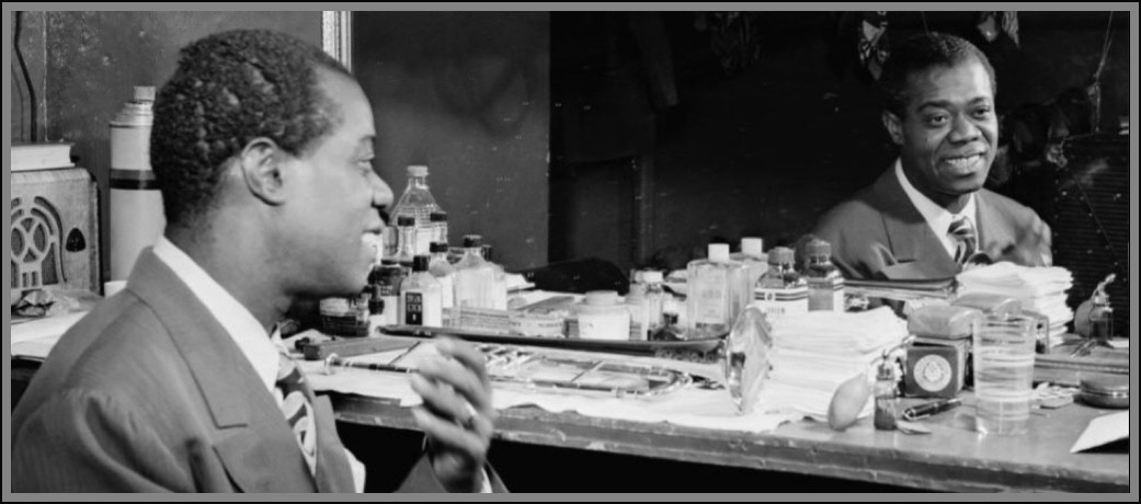 A black and white photograph by William P. Gottlieb of a young man Louis Armstrong looking into a horizontal rectangular dressing room mirror with his mirror image in mirror on right side of photograph.