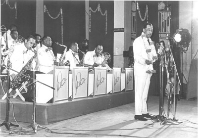 A black and white photograph of the Duke Ellington orchestra in Bombay India at the United States embassy on October 9–10, 1963 with Duke standing behind standup microphones 🎙 on the right in the photo and his orchestra sitting behind him on left.