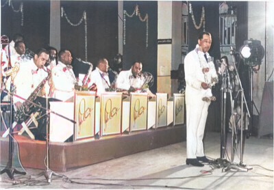 A colorized black and white photograph of the Duke Ellington orchestra in Bombay India at the United States embassy on October 9–10, 1963 with Duke standing behind standup microphones 🎙 on the right in the photo and his orchestra sitting behind him on left.