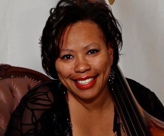 A color photograph of Nedra Wheeler in a black blouse with see through arms and plunging necklace with her bass resting against her neck on her left side.