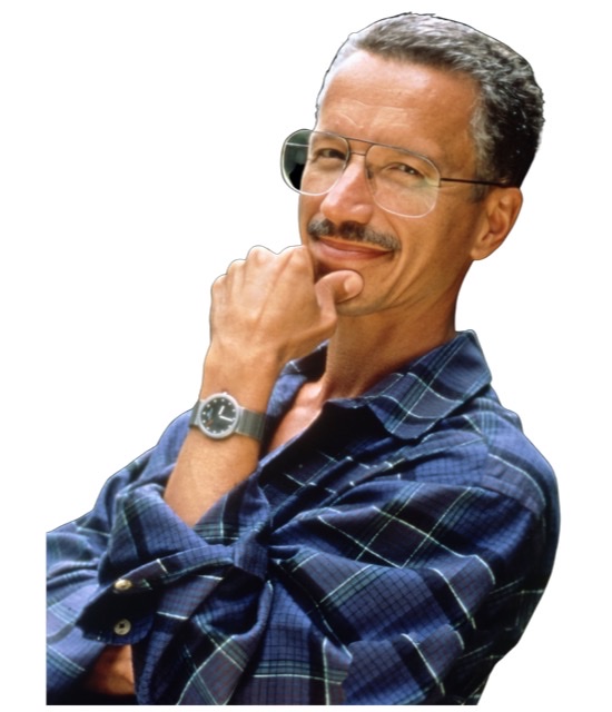 A slightly enhanced color photograph of a standing Keith Jarrett wearing glasses and  blue plaid shirt with close cropped hair and a slight smile on his face with his right hand and thumb prominently gripping his chin.