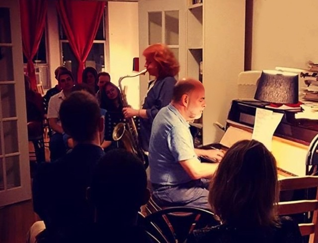 A color photograph of Lena Bloch playing her saxophone accompanied by pianist Charles Sibirsky, the owner and founder of Slope Music, in 2018 surrounded by a small audience sitting in chairs looking in from multiple rooms.