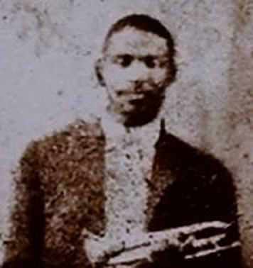 A grainy closeup of a black and white photograph of Buddy Bolden's head and shoulders holding trumpet in lower right corner of picture.