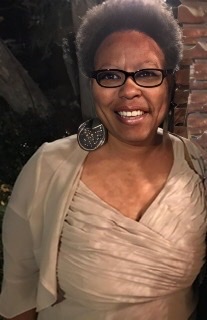 A color photograph of Nedra Wheeler wearing black framed glasses in a full-length beige puffy dress with a matching beige jacket.