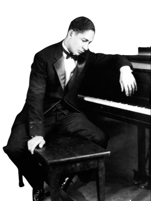 Jelly Roll Morton as a young man in a tuxedo sitting on a piano bench draped over the front of a piano with head bowed.