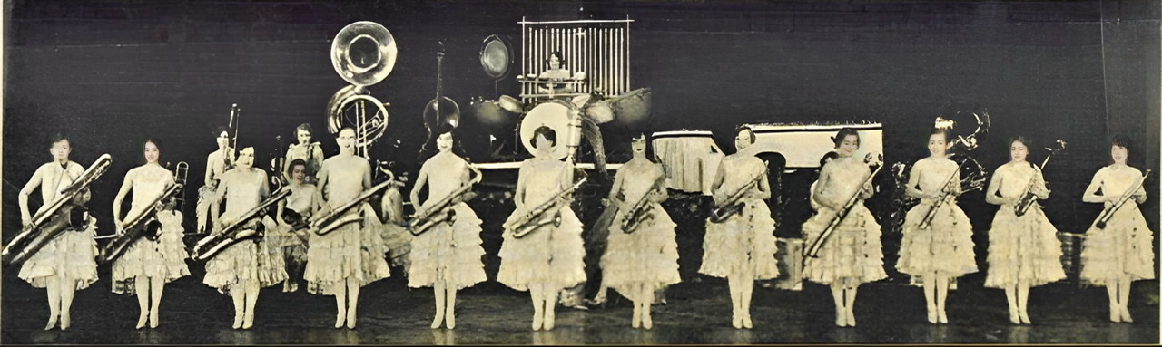 An enhanced black and white photograph from a German newspaper of The Ingenues lined up across the stage facing the audience holding twelve saxophones of various types with other band members in the background.