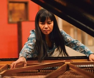 An enhanced color photograph of Satoko Fujii  wearing a silky blue shirt with brown designs on it reaching into her piano viewed from inside of the piano.