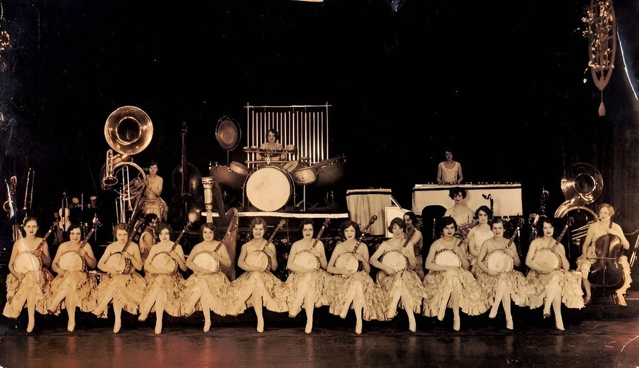 An enhanced photograph of the twenty-member orchestra with twelve sitting, cross-legged members with banjos 🪕 lined up across center stage.