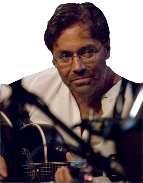 A color photograph of a closeup mostly headshot of Al Dimeola with open framed glasses wearing an multiply unbuttoned low profile collar white shirt sitting on stage playing his dark blue/purple acoustic guitar with his eyes turned to his left.