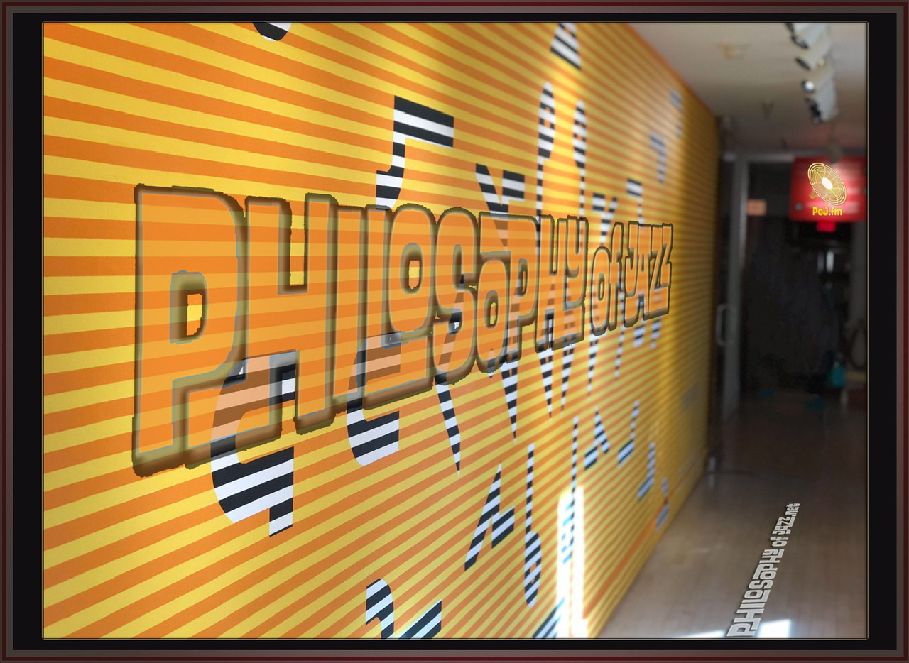 A framed photograph if a yellow metal corrugated wall running down the left side of a corridor with Philosophy of Jazz written on it and a PoJ.fm logo as the emergency light towards end of corridor on the right.